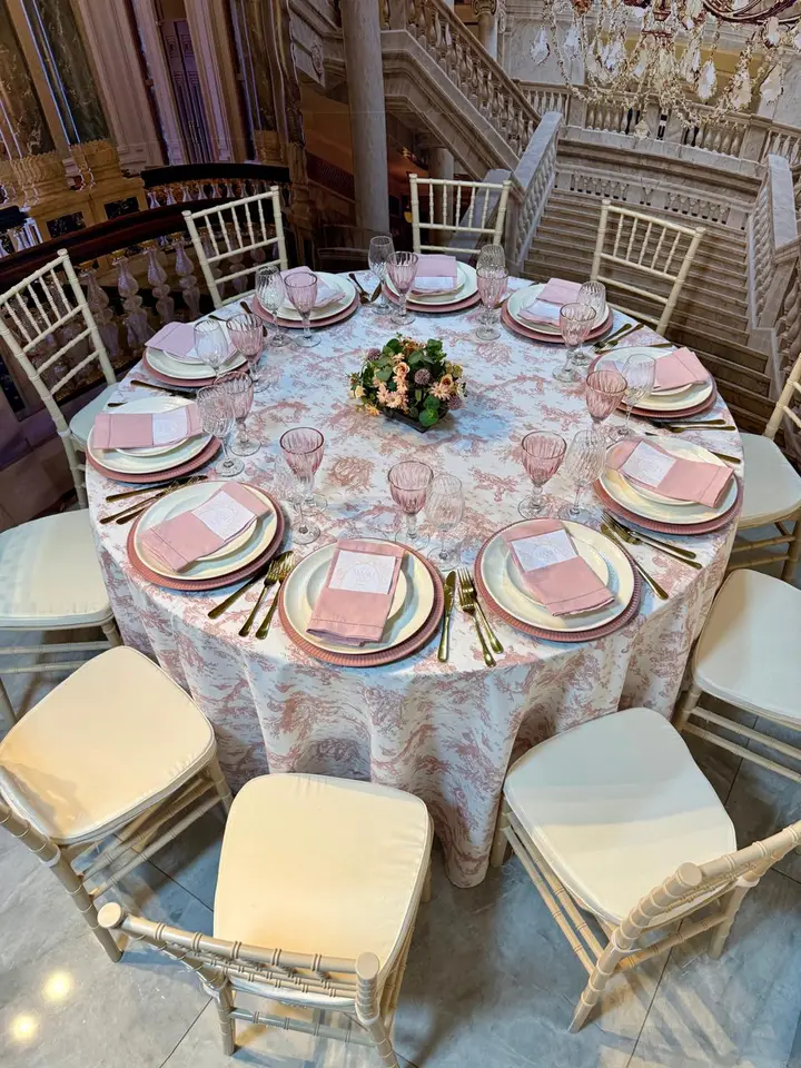 Mesa redonda posta em salão, com toalha floral, guardanapos rosa e centro de mesa