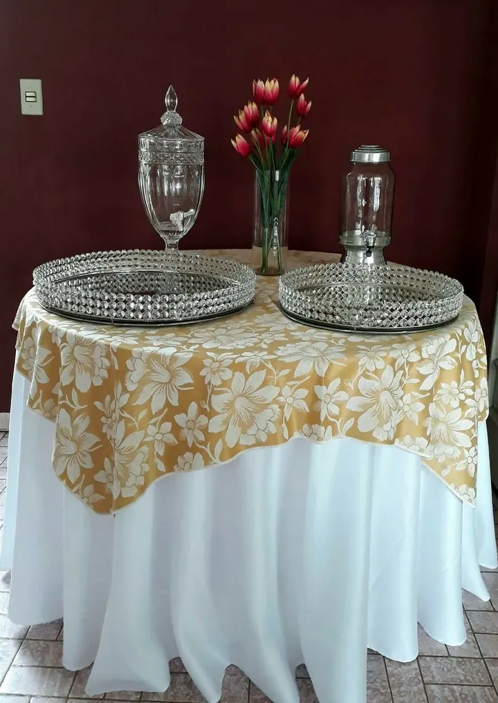 Mesa redonda com toalha branca e sobretoalha floral dourada, taças de cristal e flores